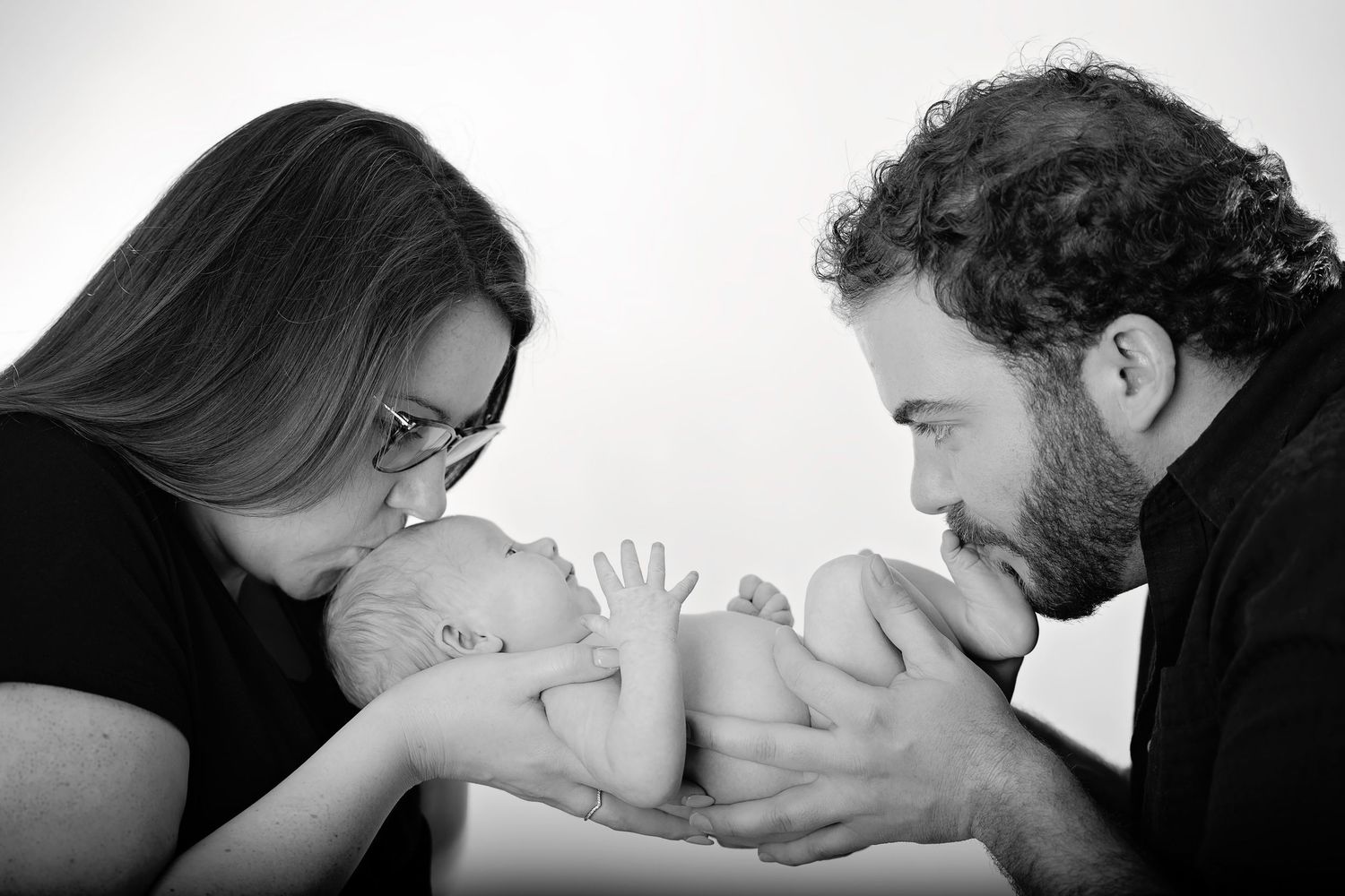 Black and white portrait of parents lovingly kissing their newborn baby together.