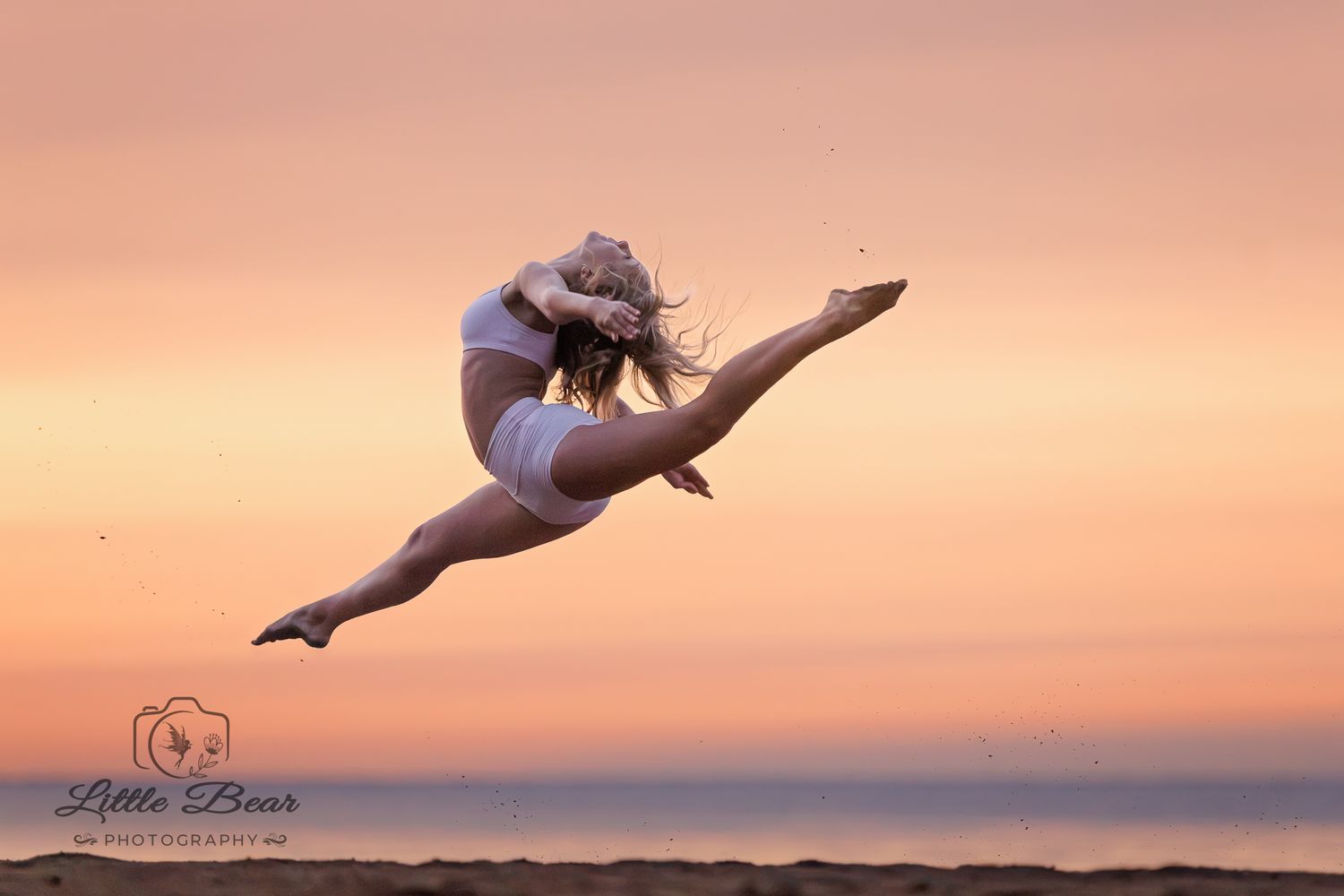 Dancer leaping gracefully at sunset on a beach, performing a split jump against a pink and orange sky.