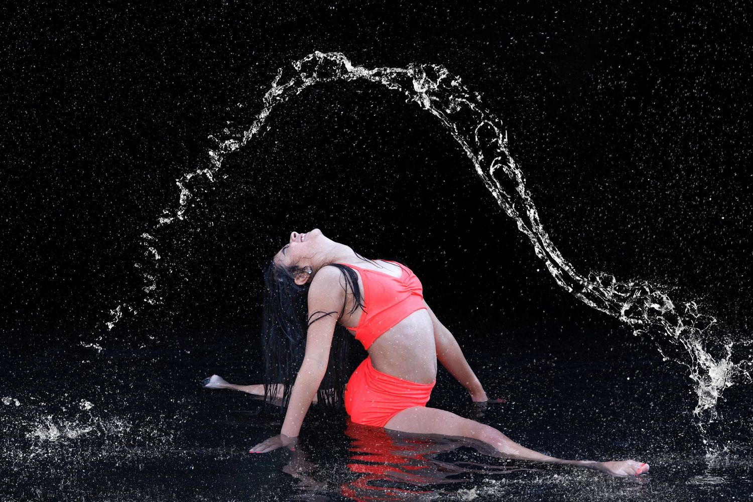 A dancer in a red outfit kneels dramatically as a water arc splashes around her against a dark background.