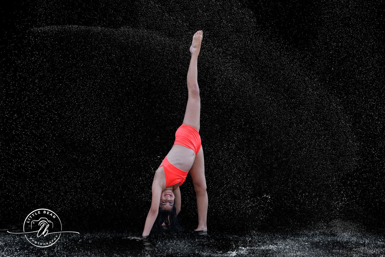Gymnast in red outfit performs a backbend on wet floor with dramatic water spray against black background.