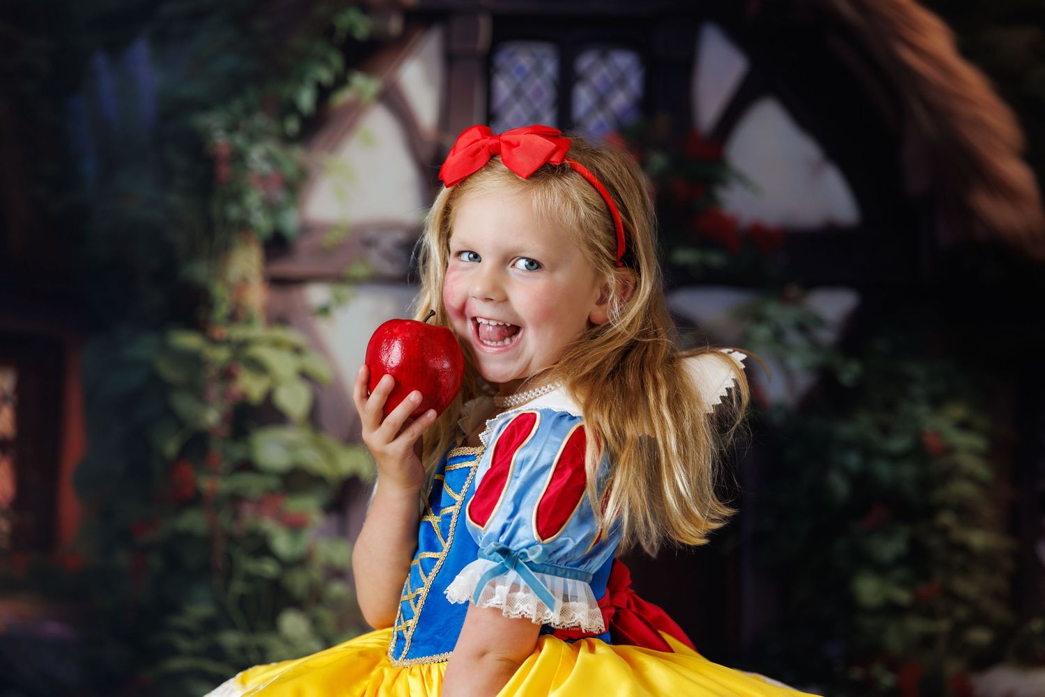 Young girl dressed as Snow White holding a red apple, wearing blue and yellow costume with red bow headband.
