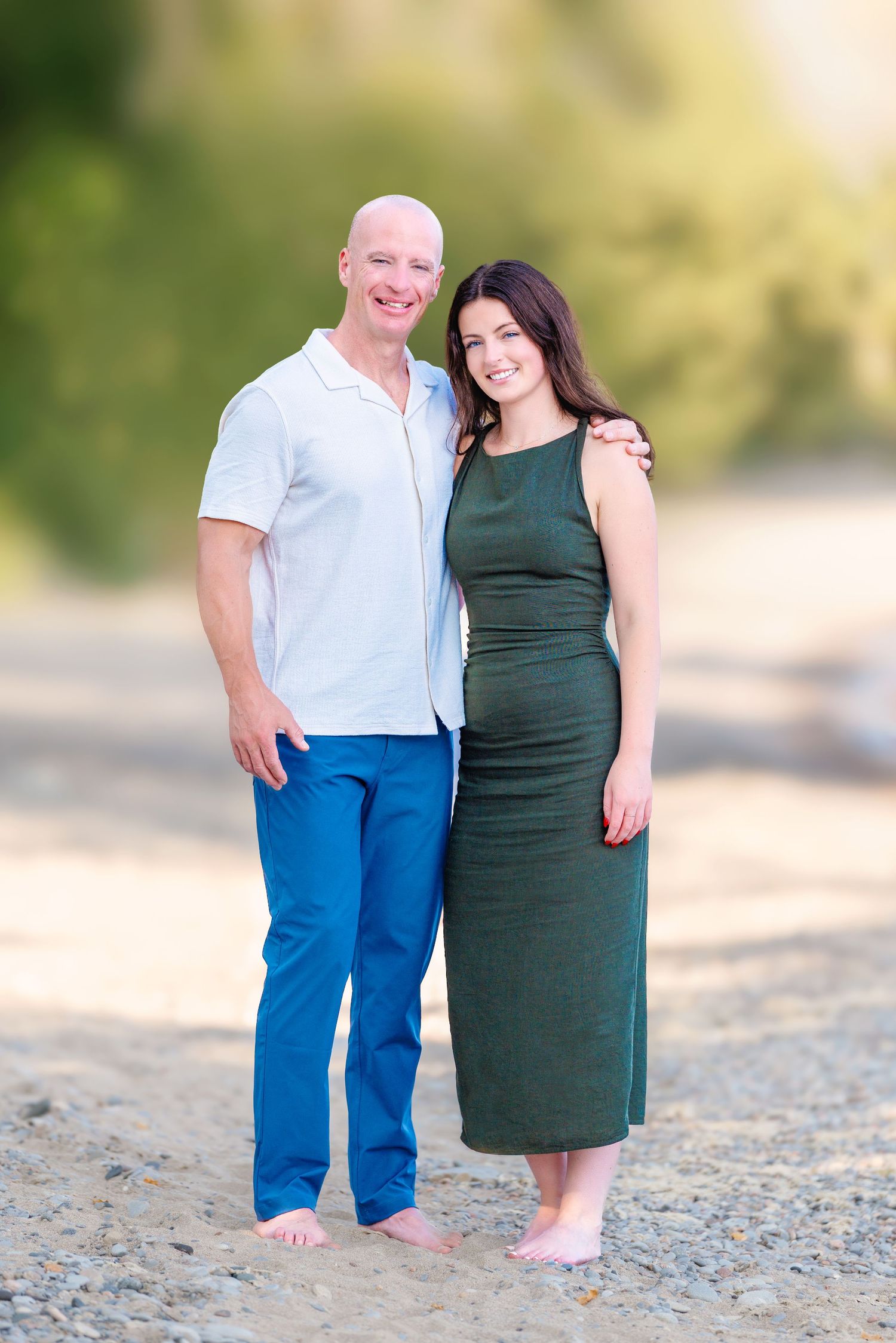 Bald man in white shirt and blue pants poses with brunette woman in green dress on a sandy beach.