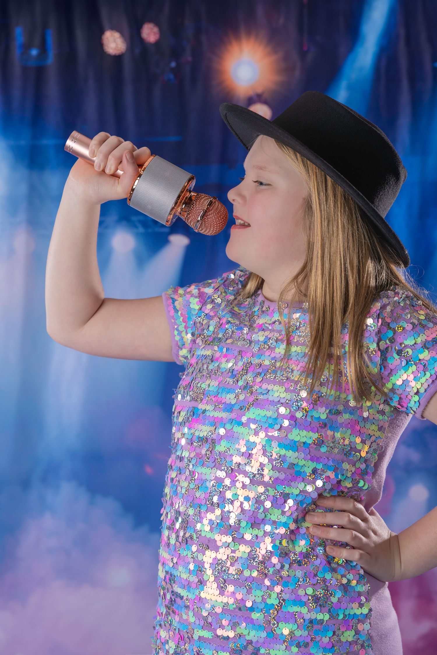 Young girl in sequin dress and black hat singing into microphone on a dramatic lit stage.