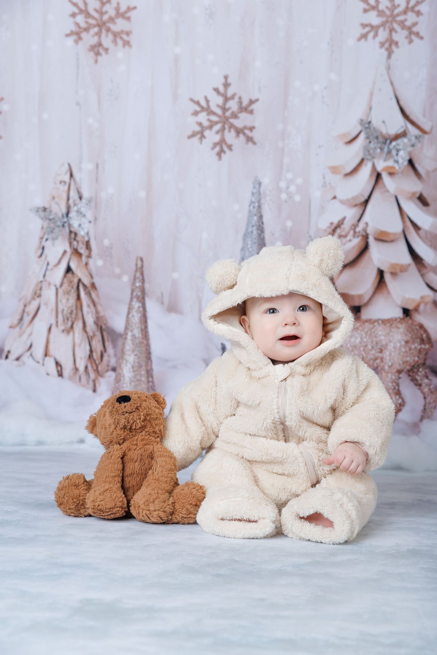 Adorable baby in cream bear costume sits with teddy bear in a winter wonderland studio setting.