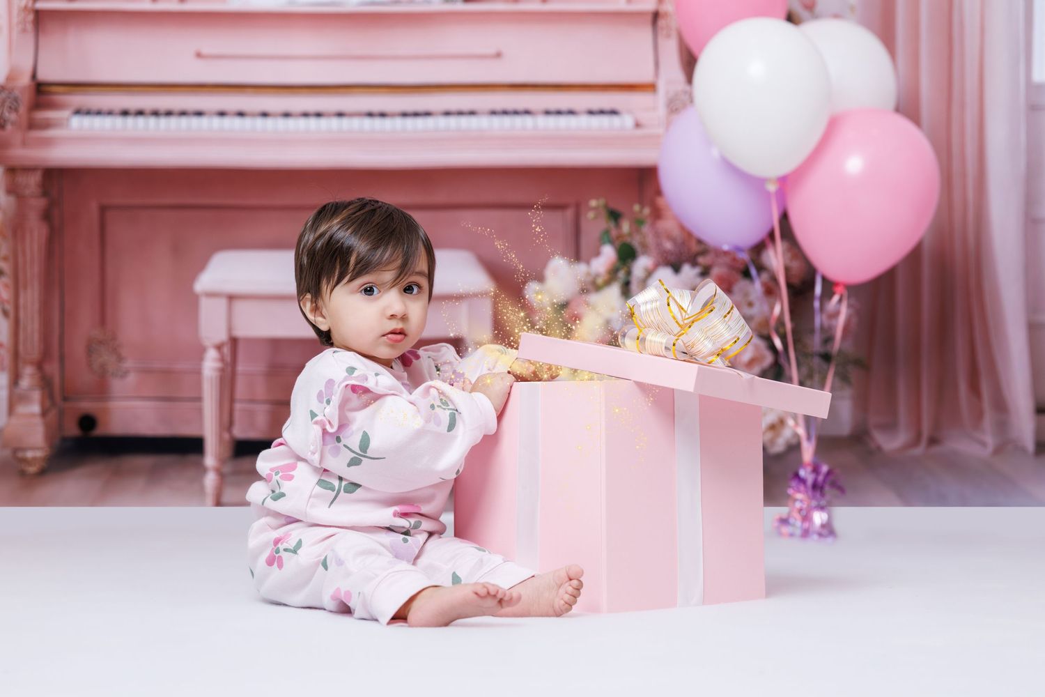 A baby girl in floral outfit reaches into a pink gift box in a decorated studio setting.