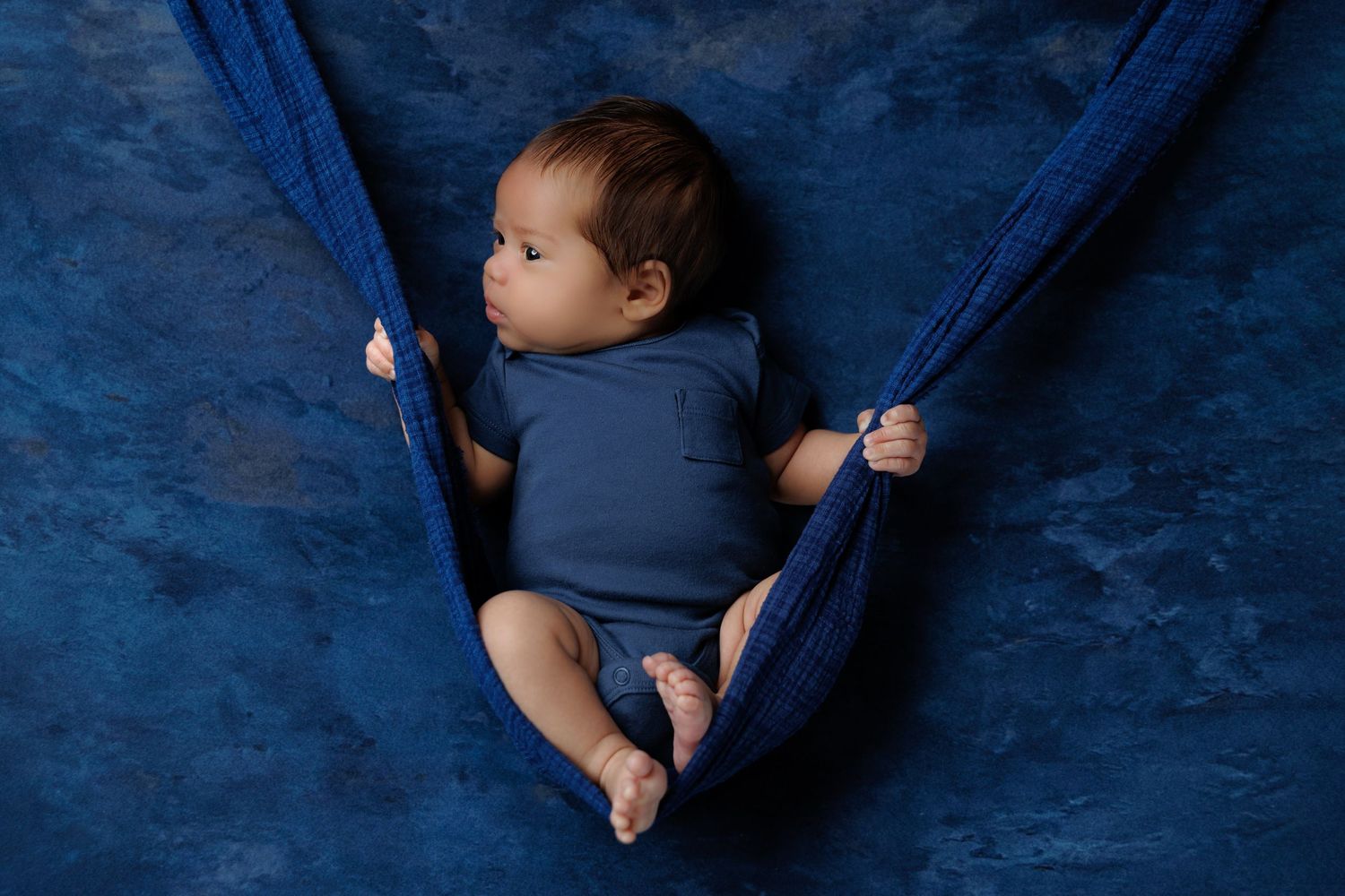 Baby in blue onesie lies in fabric hammock against deep blue textured background.