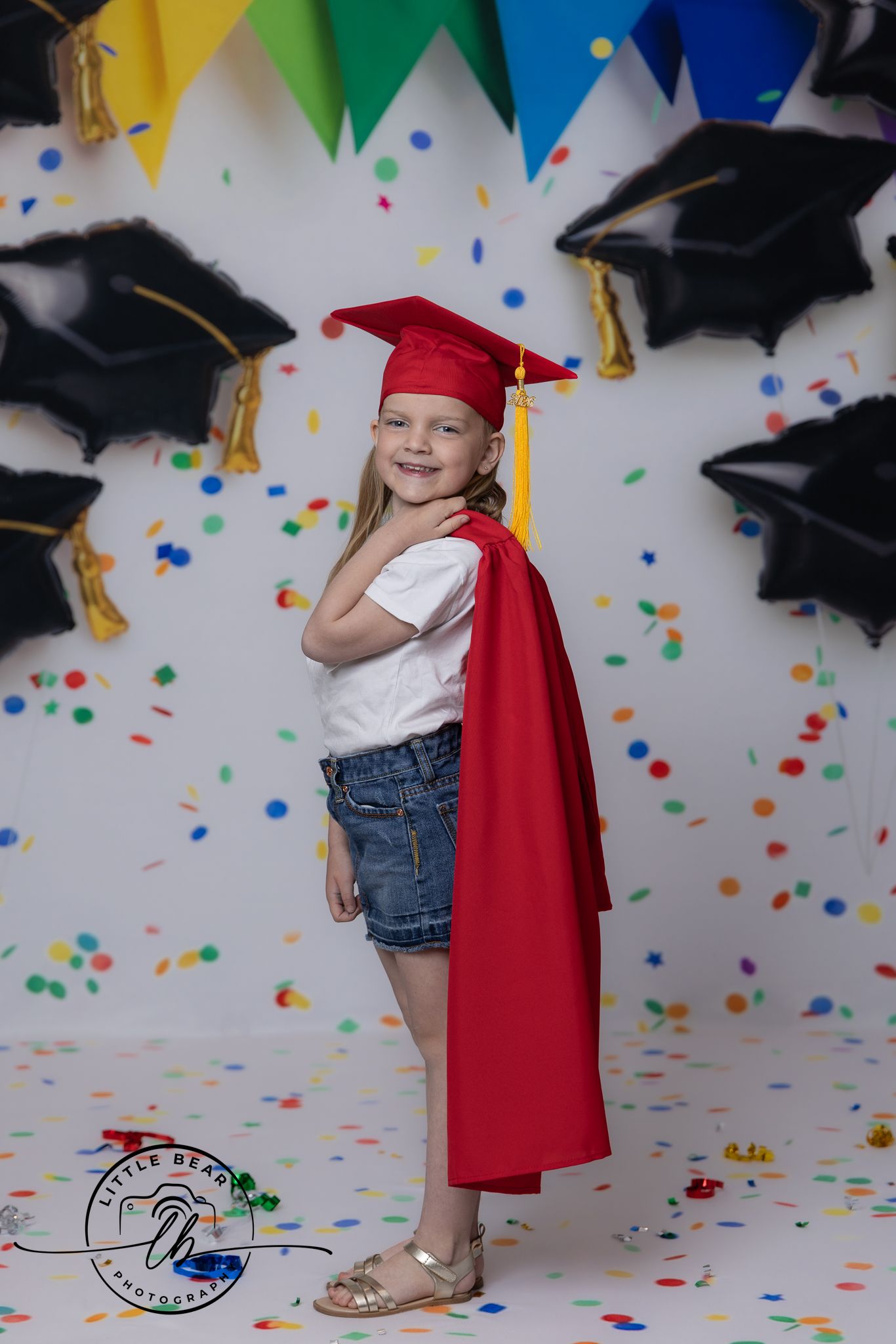 Young girl in red graduation cap and gown smiles proudly among confetti and grad balloons at her preschool graduation.