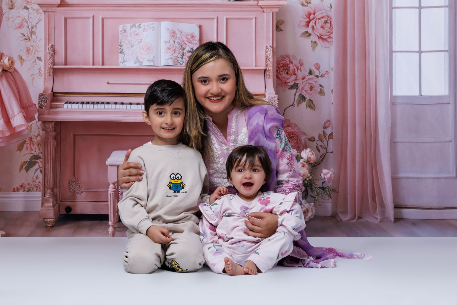 Mother smiles with young son and baby daughter seated in front of pink piano in floral studio.