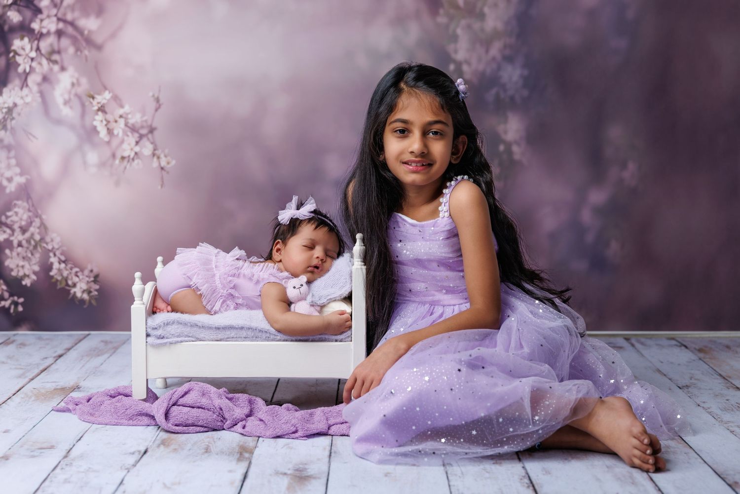 Young girl in purple dress sits beside newborn sibling in miniature bed in studio portrait.