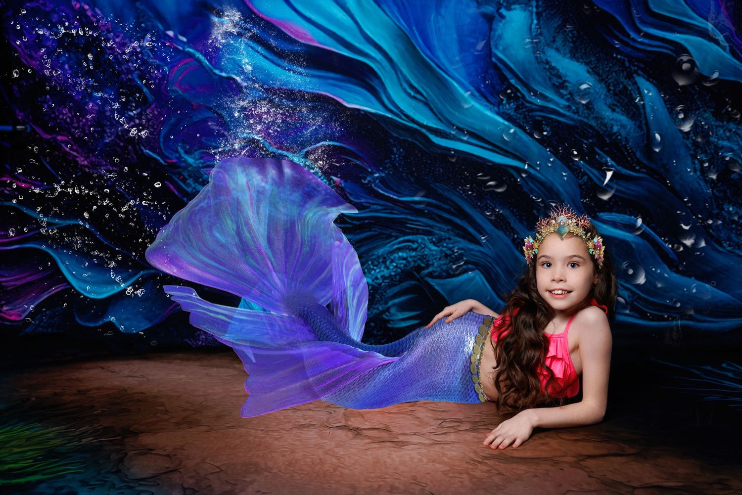 Young girl in red and purple mermaid costume poses lying down against swirling blue ocean backdrop.