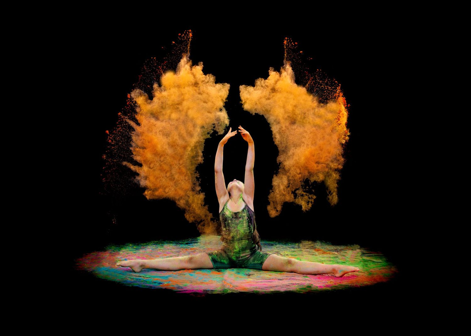 Dancer in green leotard performs graceful pose surrounded by vibrant orange color powder explosion on black background.