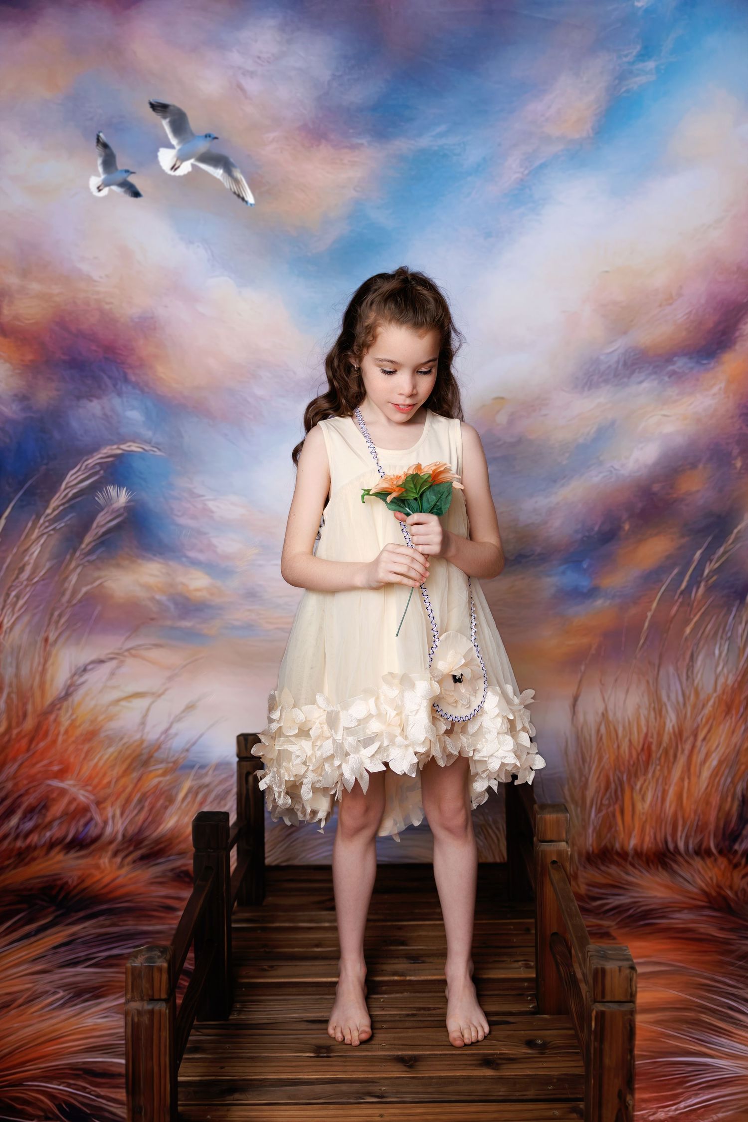 A young girl in a cream petal-hem dress holds orange flowers while standing on a wooden bridge with a sky backdrop.