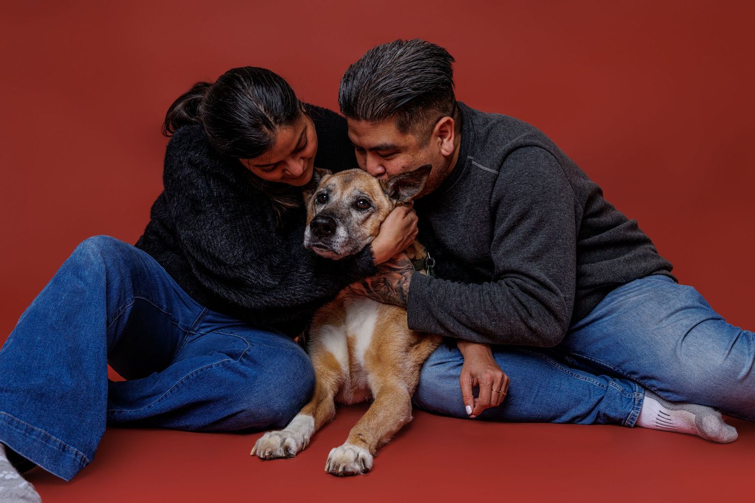 A couple sitting on a red studio backdrop, affectionately petting their brown mixed-breed dog between them.