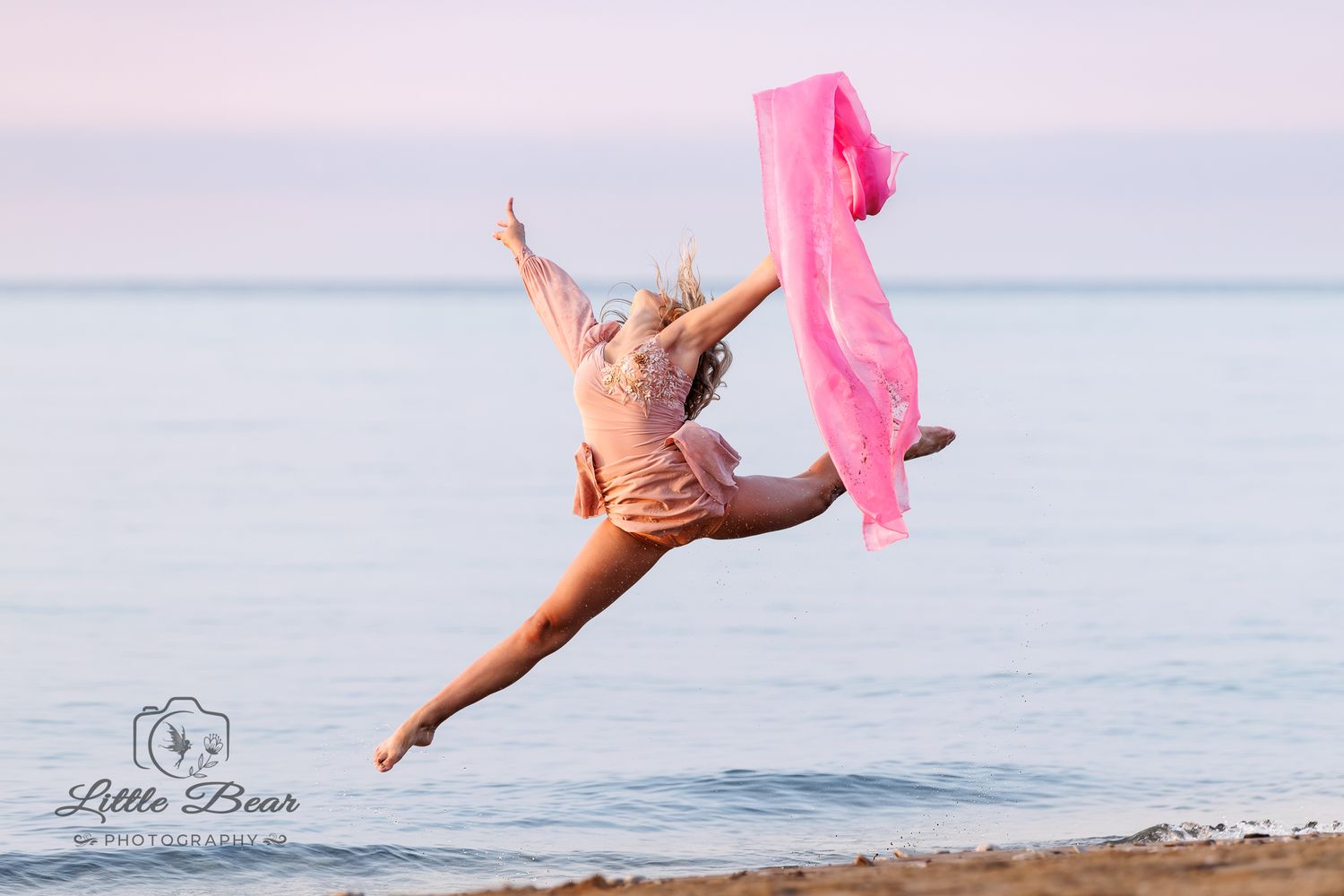 Young dancer leaps gracefully on a beach at sunset, holding a flowing pink fabric scarf against a pastel sky.