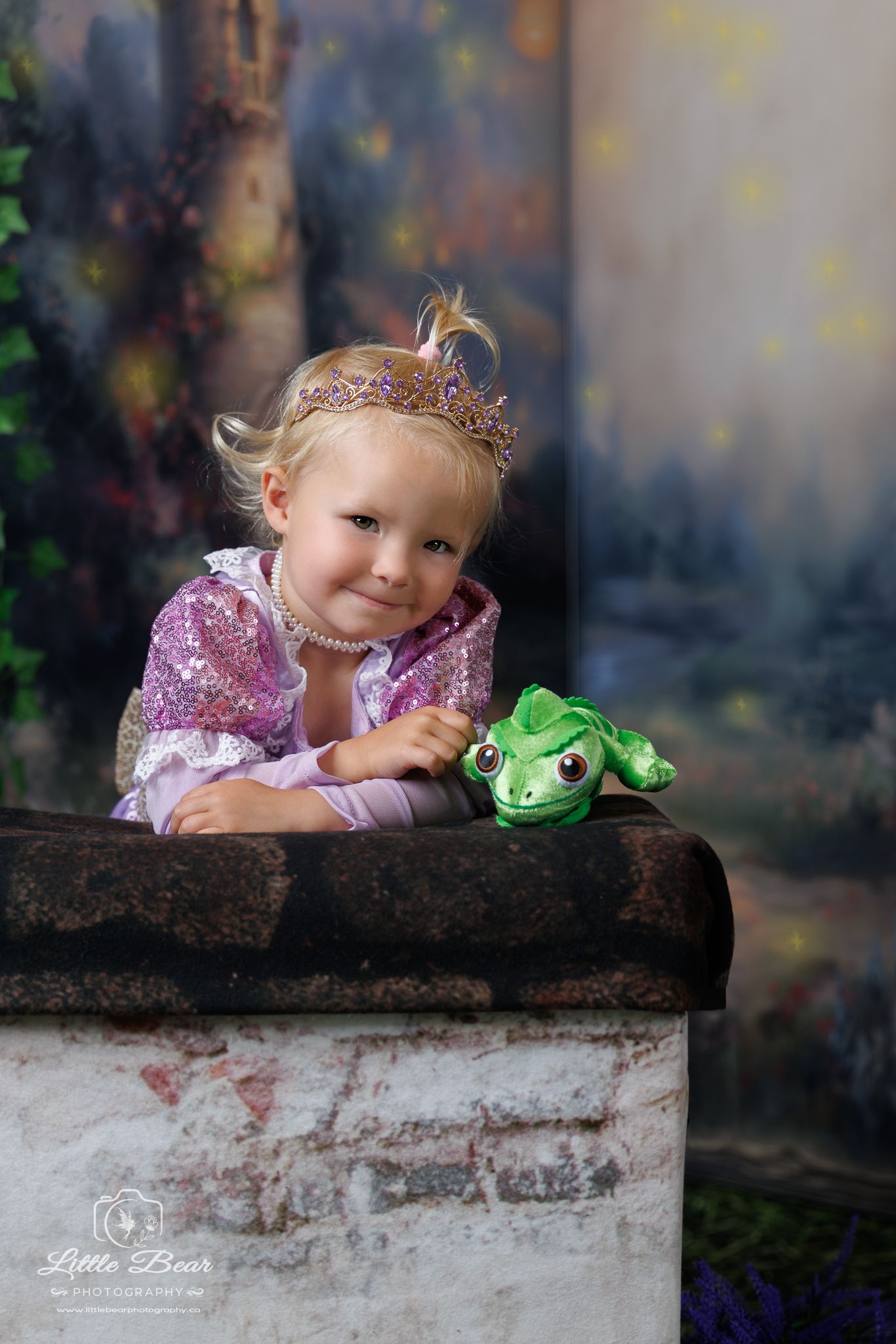 Toddler girl dressed as Rapunzel with crown and pearl necklace holding Pascal frog stuffed animal on stone wall.