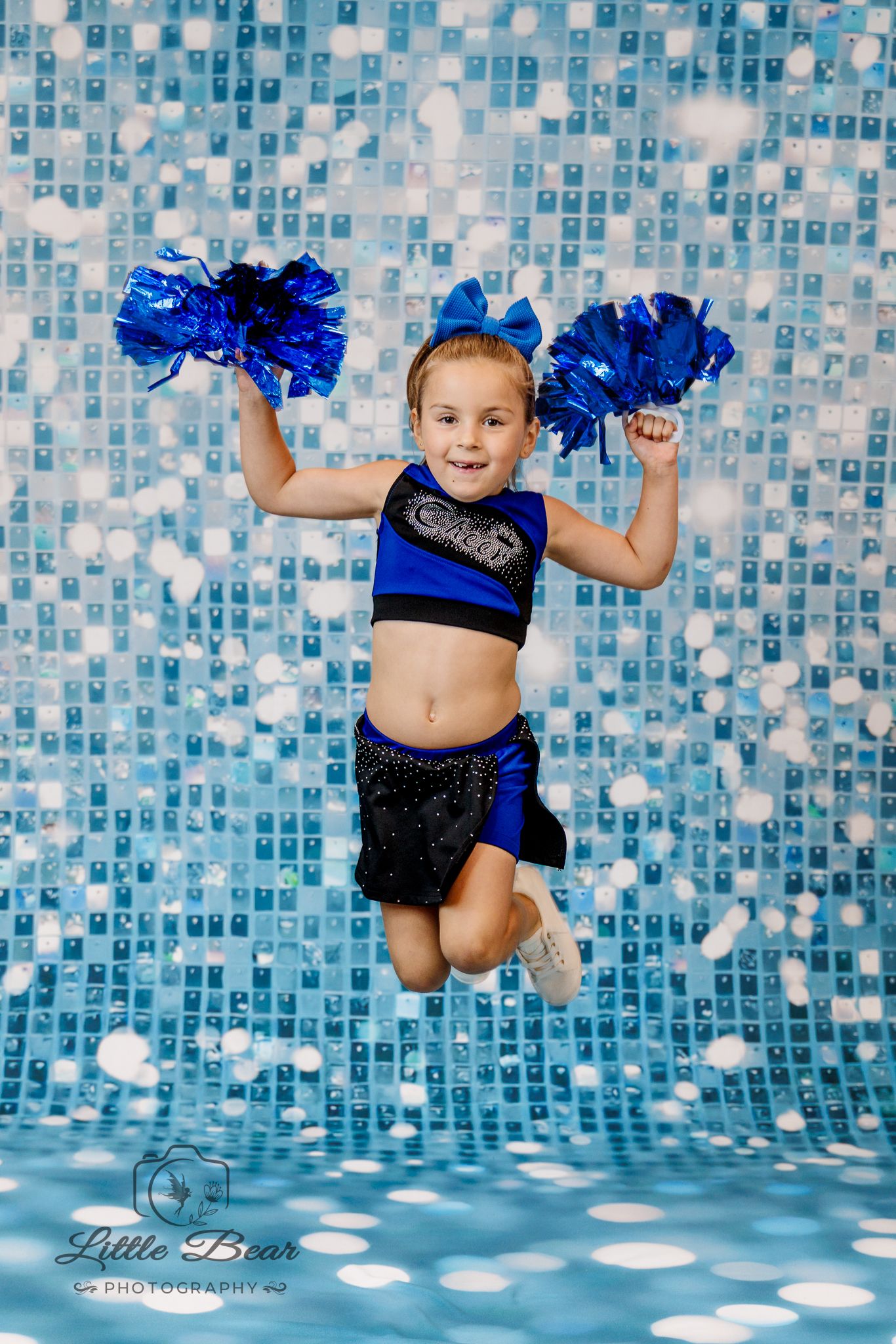 Young girl in blue cheerleader uniform jumping with blue pom-poms in front of sparkly blue backdrop.