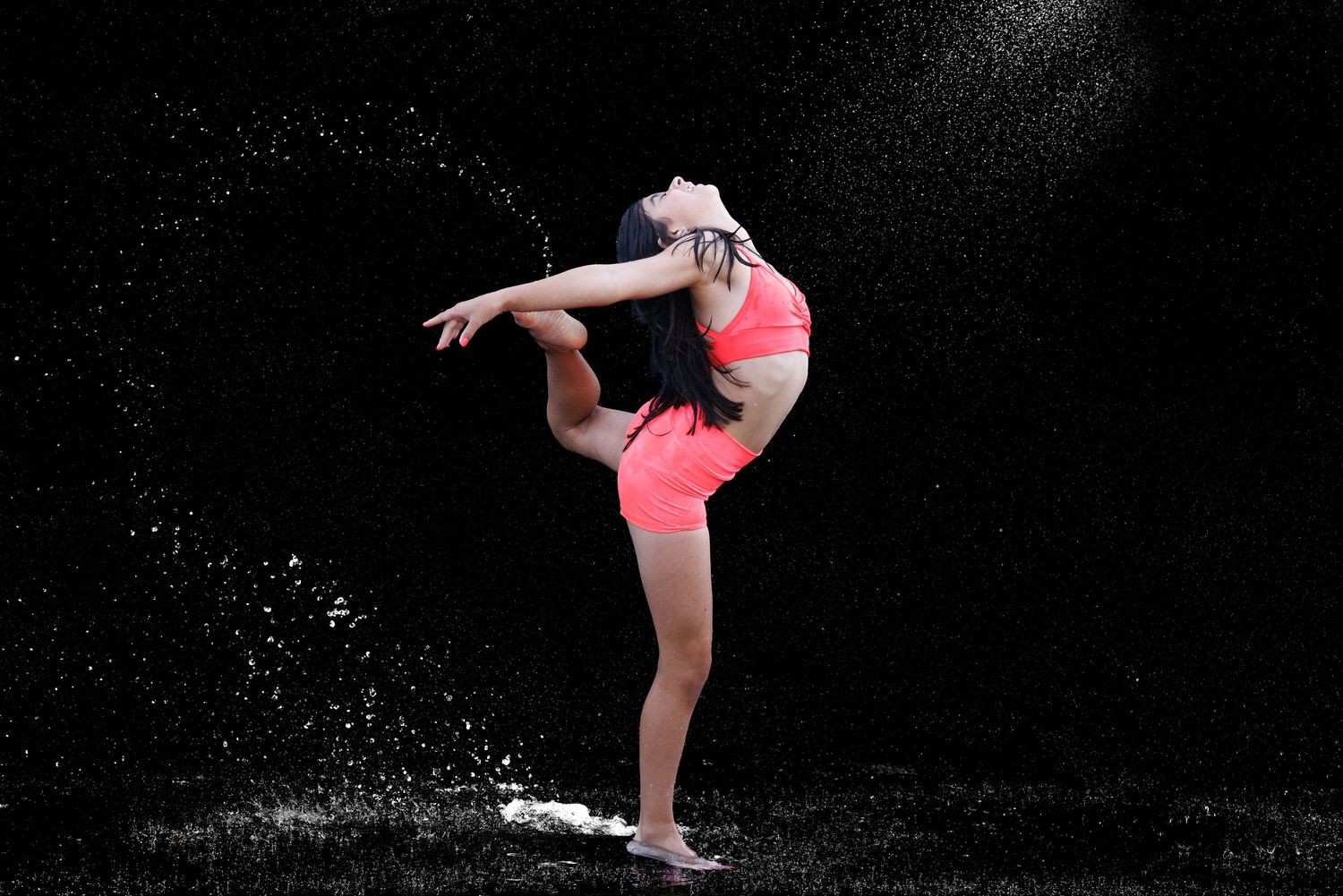 A female dancer in pink athletic wear performs a graceful high kick pose with water droplets splashing around her on a dark background.