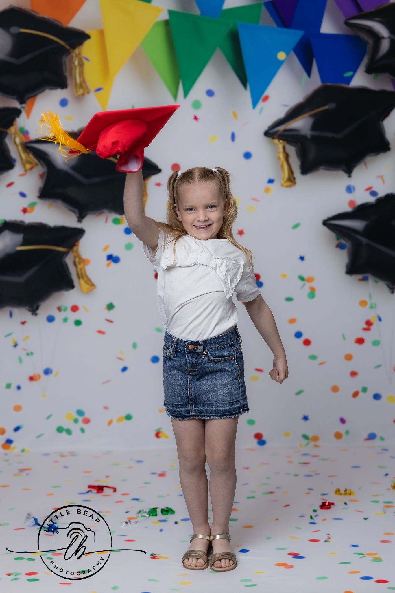 Young girl in white top and denim skirt celebrating graduation, holding red cap with confetti and mortarboard balloons.
