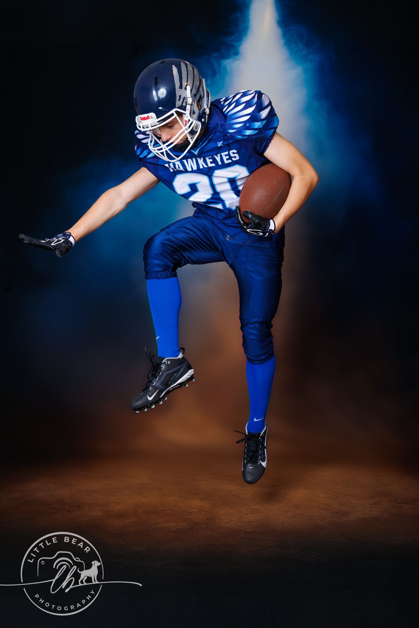 Young Hawkeyes football player #20 jumping mid-air with ball in blue uniform against dark smoky background.