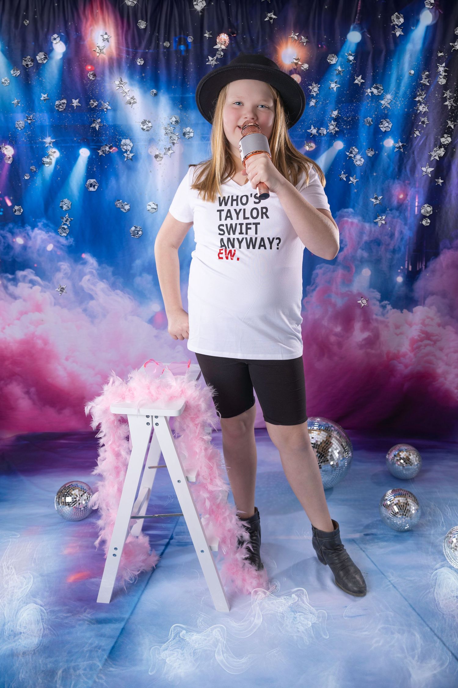 Young blonde girl in Taylor Swift tee and black hat sings into microphone surrounded by disco balls on stage.
