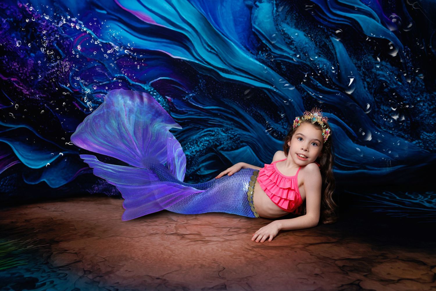 Young girl in pink top and purple mermaid tail lies on floor against vibrant blue painted backdrop.