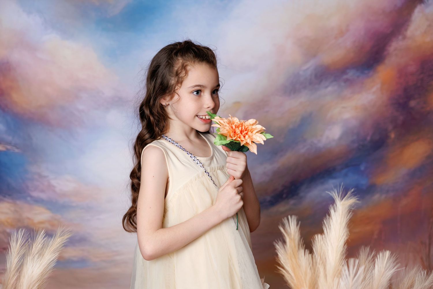 A smiling young girl in a cream dress holds an orange flower against a vibrant painted sky and pampas grass backdrop.