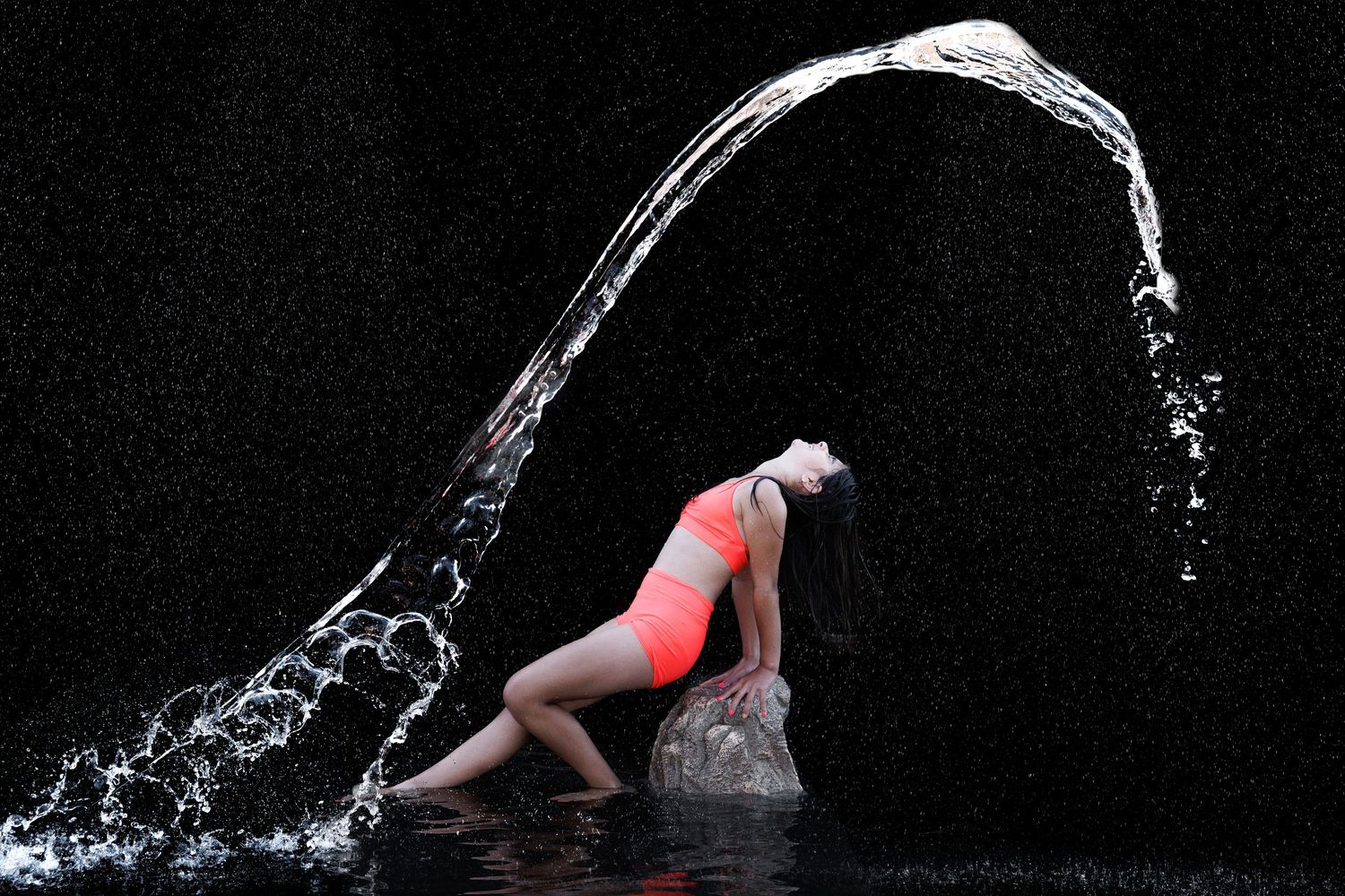 Woman in pink bikini poses on rocks as a dramatic arc of water splashes around her against a dark background.