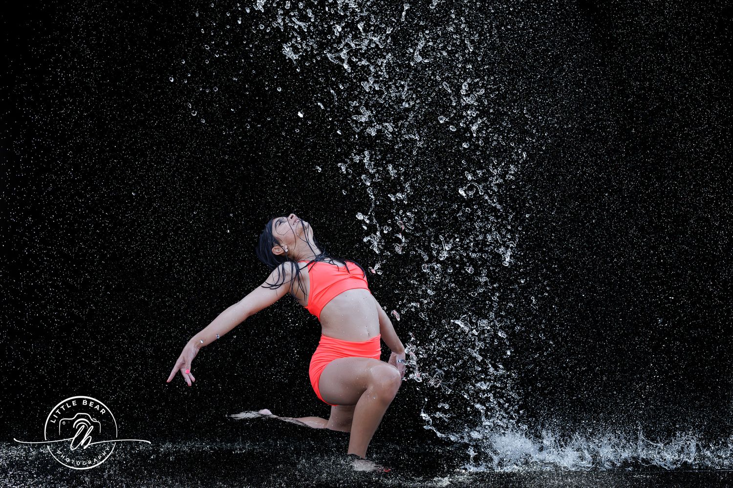 Dancer in red bikini poses gracefully amid splashing water against dark background.