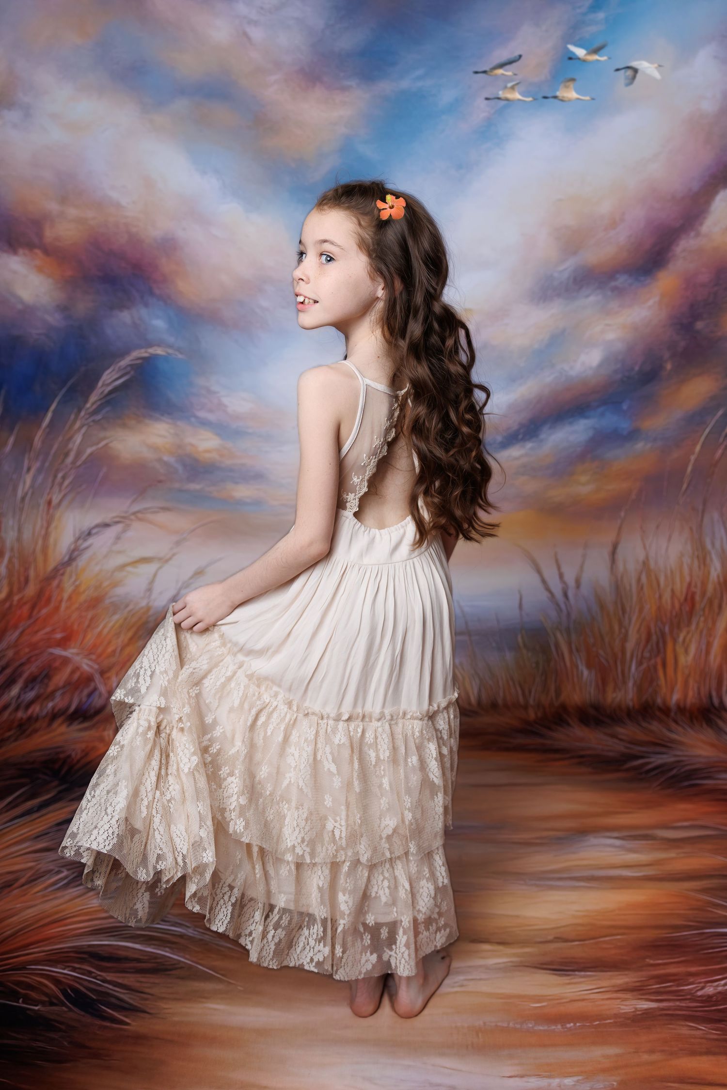 A girl in a backless cream lace dress poses gracefully before a painted sunset and tall grass scene.
