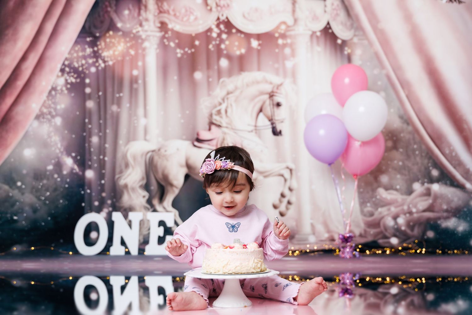 A baby girl in pink sits before a white birthday cake with ONE signs and pink balloons.