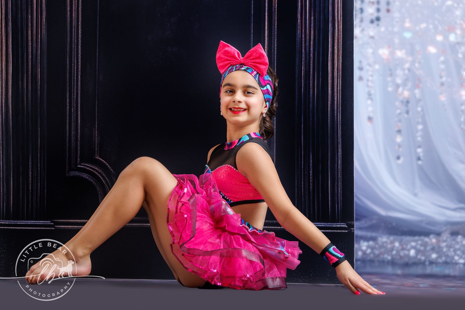 Young girl in a pink ruffled dance costume and bow headband poses gracefully on a glittery studio backdrop.