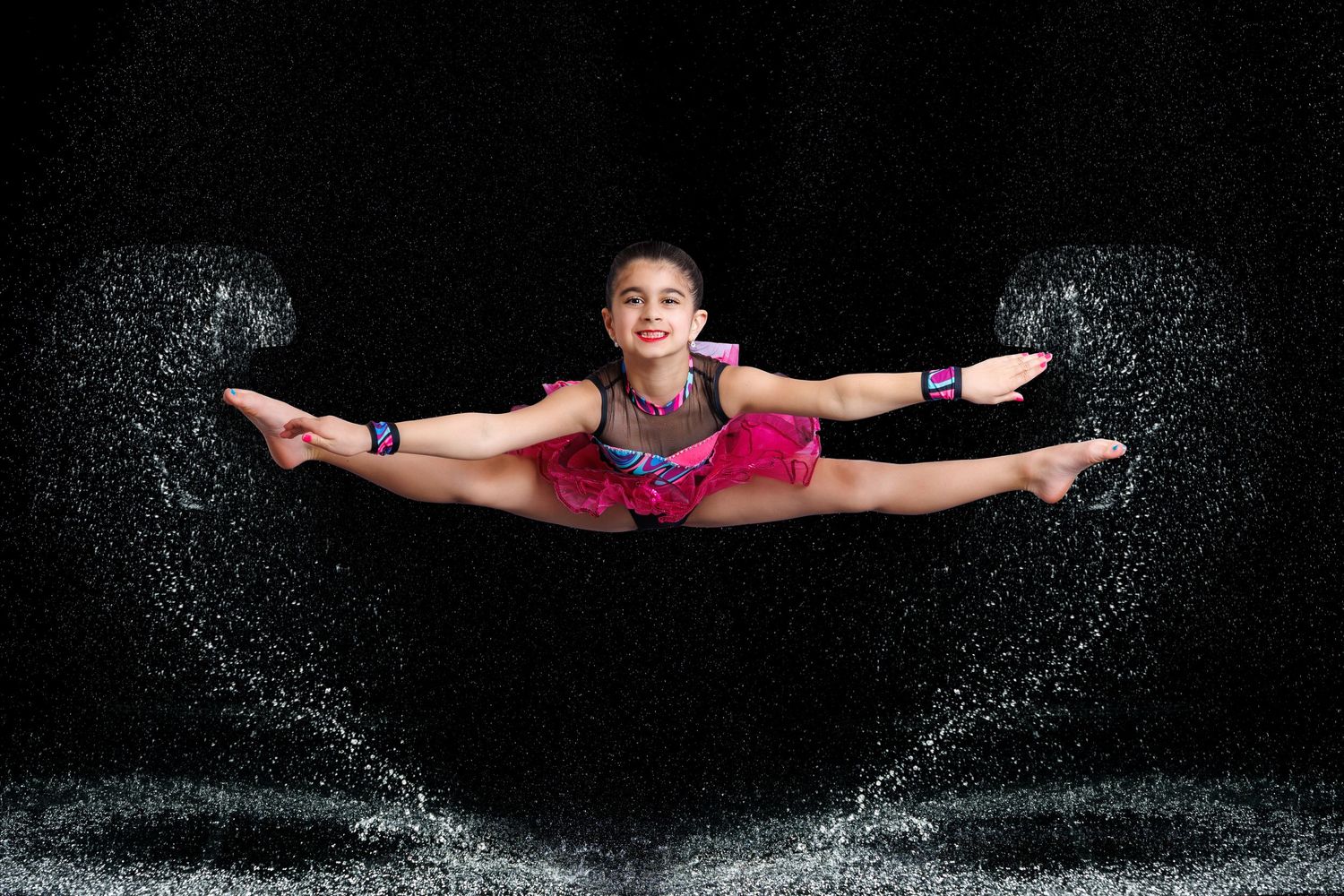 Young gymnast performs a stunning mid-air split leap in a pink costume with dramatic water splash effects on black background.