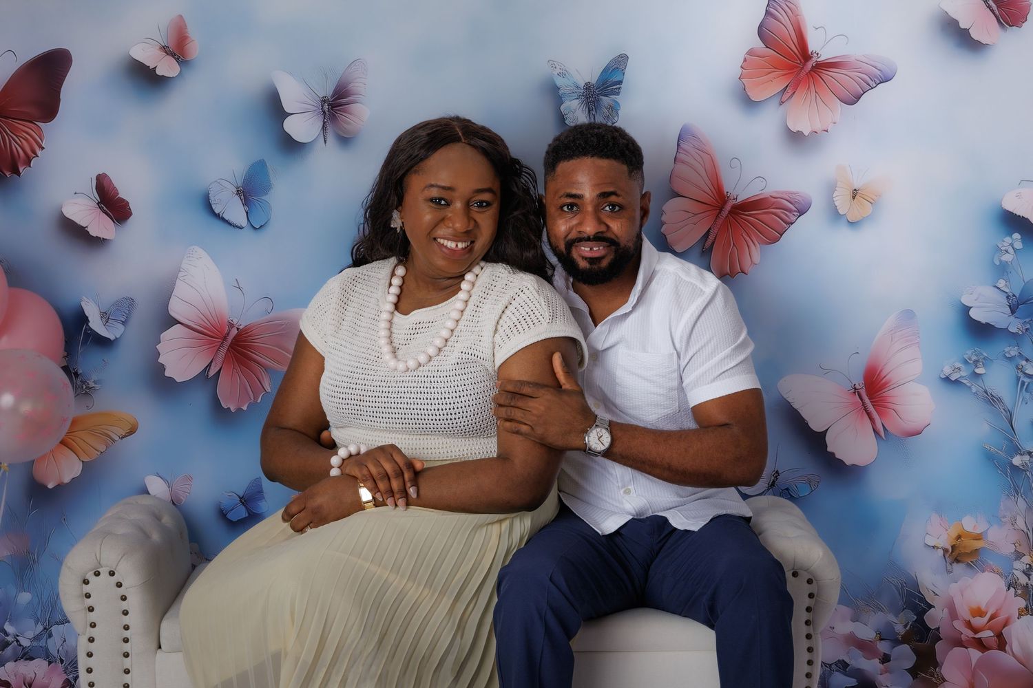 Couple smiling and posing together on a white chair against a colorful butterfly backdrop in a portrait photo.