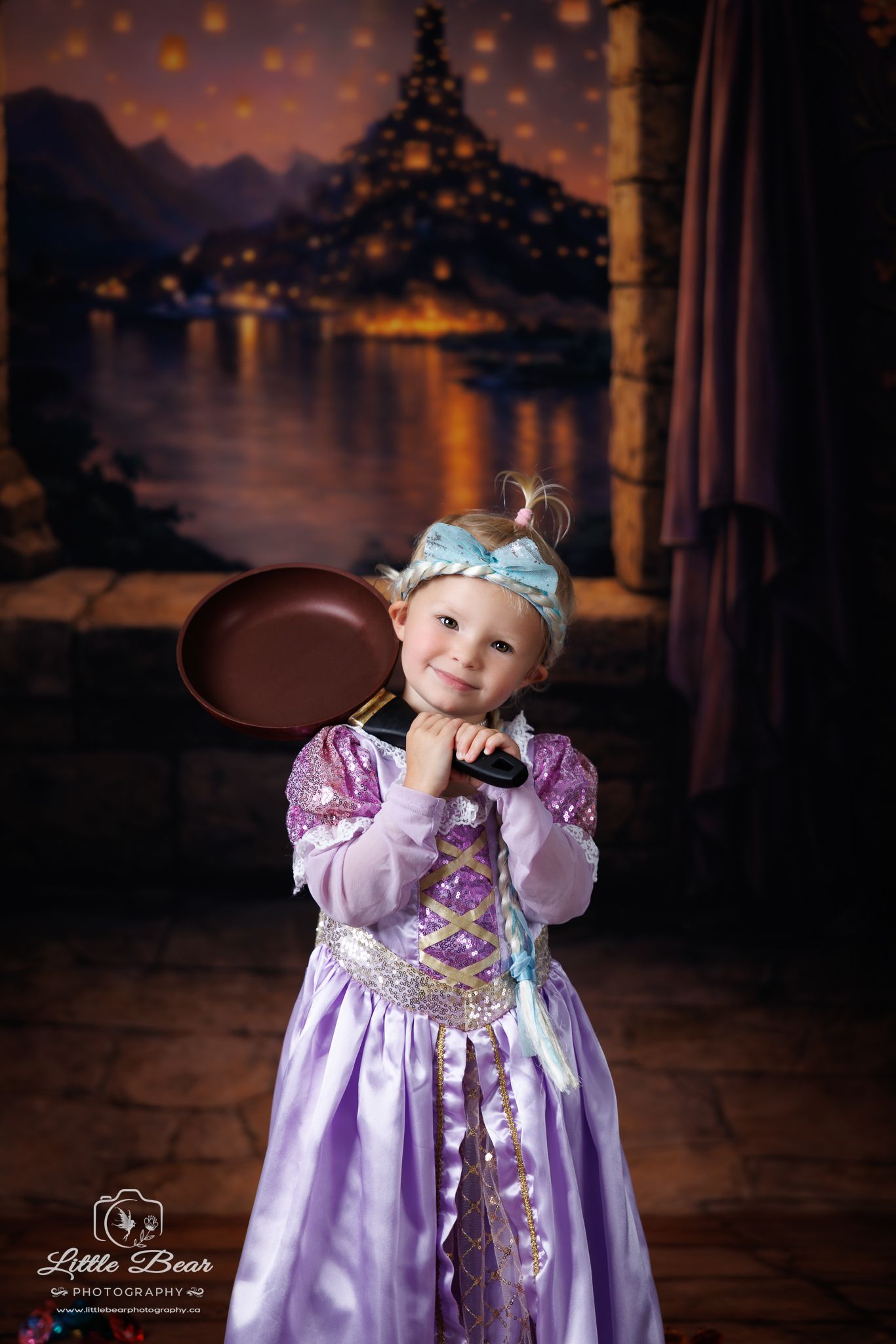 Toddler girl dressed as Rapunzel in purple princess gown holds frying pan with Tangled tower backdrop.