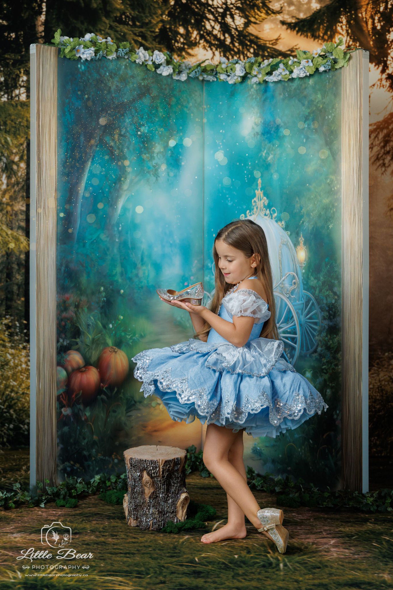 Young girl in blue Cinderella dress holds glass slipper in enchanted forest backdrop with carriage, styled fairy tale photo.