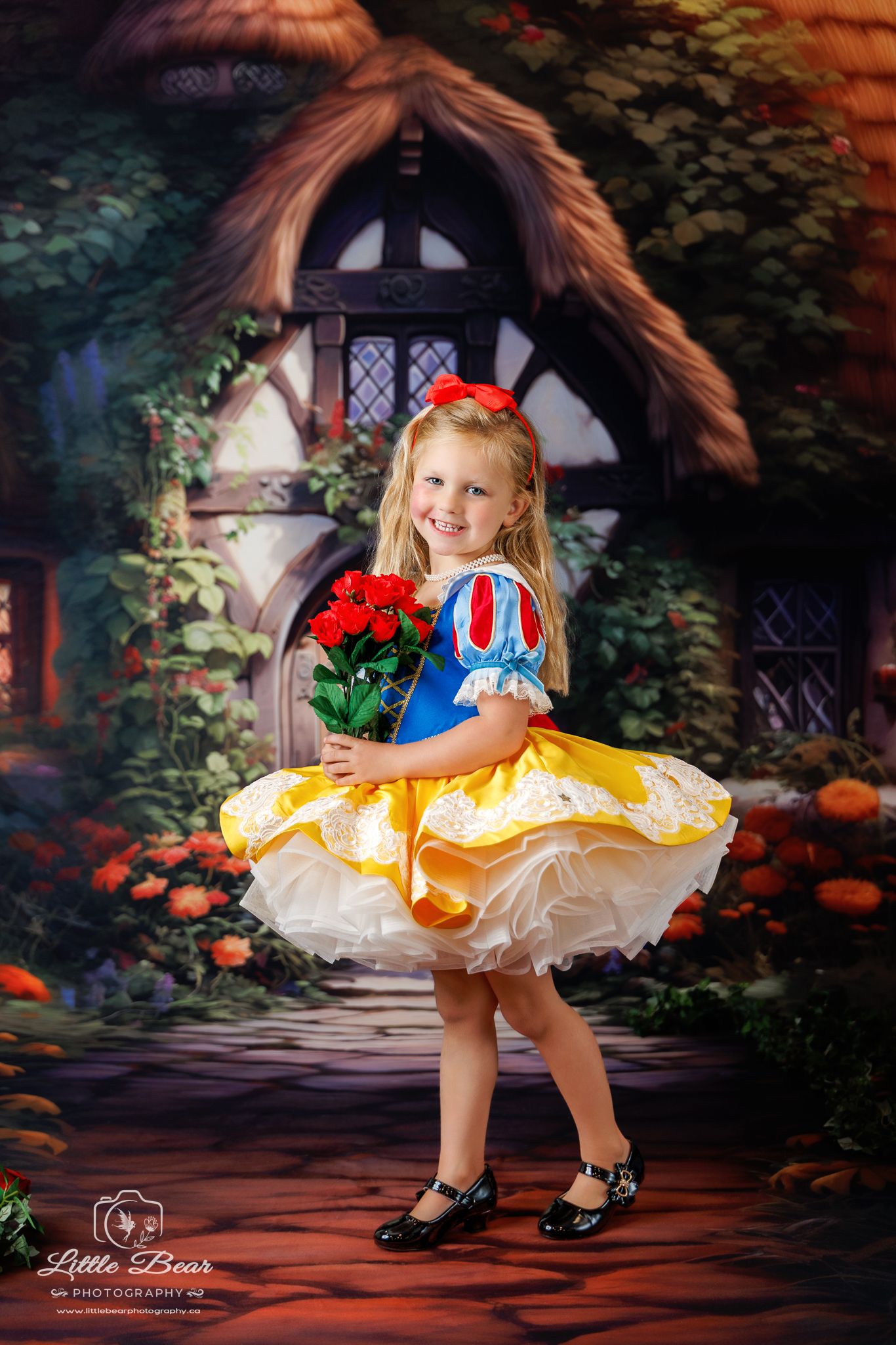 Young girl in Snow White costume holding red roses, posing in front of a fairy tale cottage backdrop.