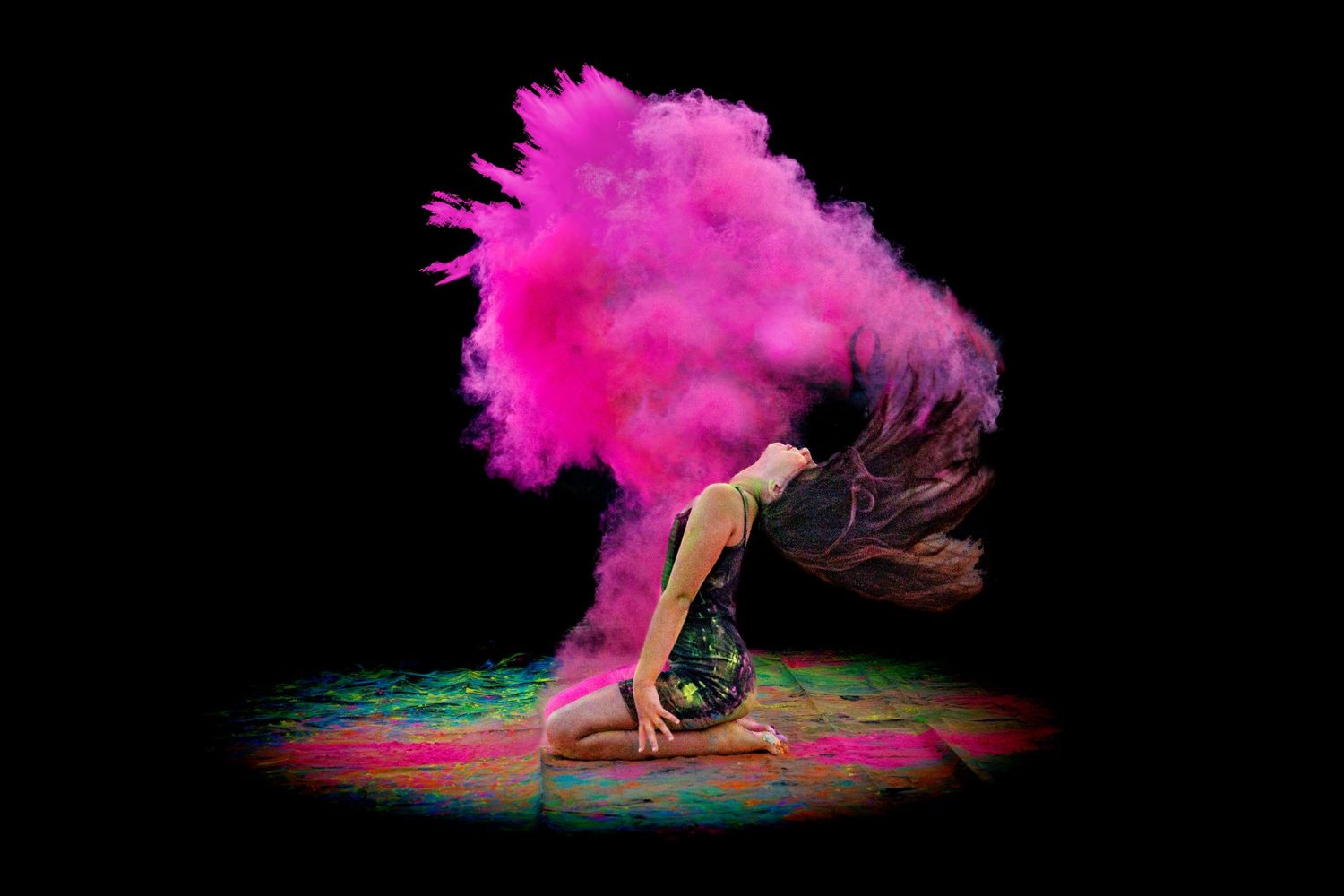 Dancer kneels dramatically as vibrant pink powder explodes around their flowing hair on dark background.