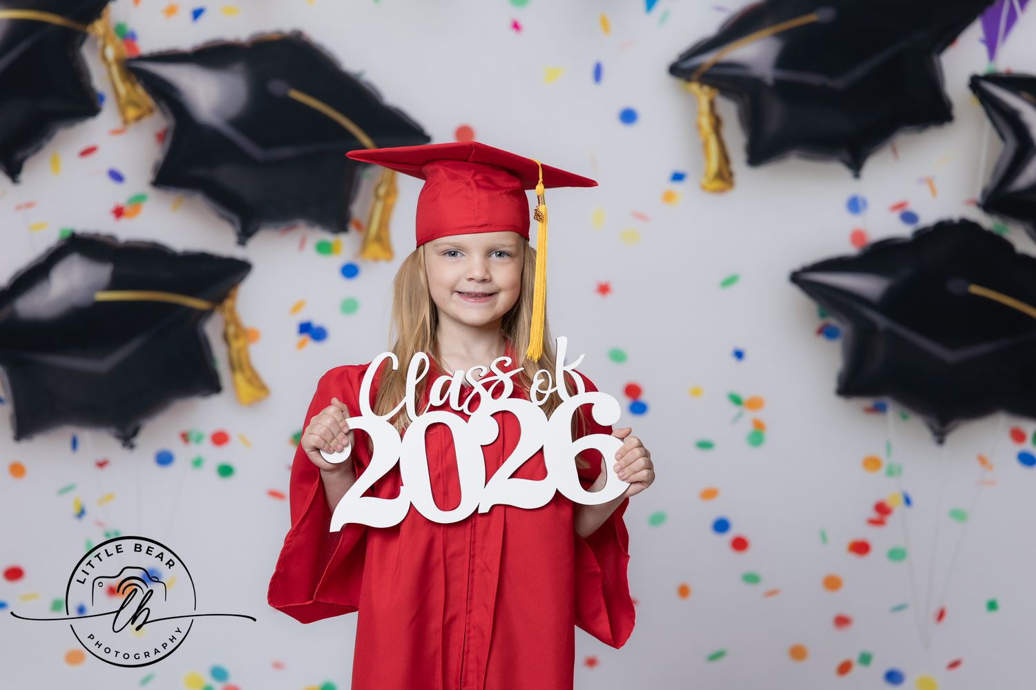 Young girl in red graduation cap and gown holds Class of 2026 sign with balloon backdrop.