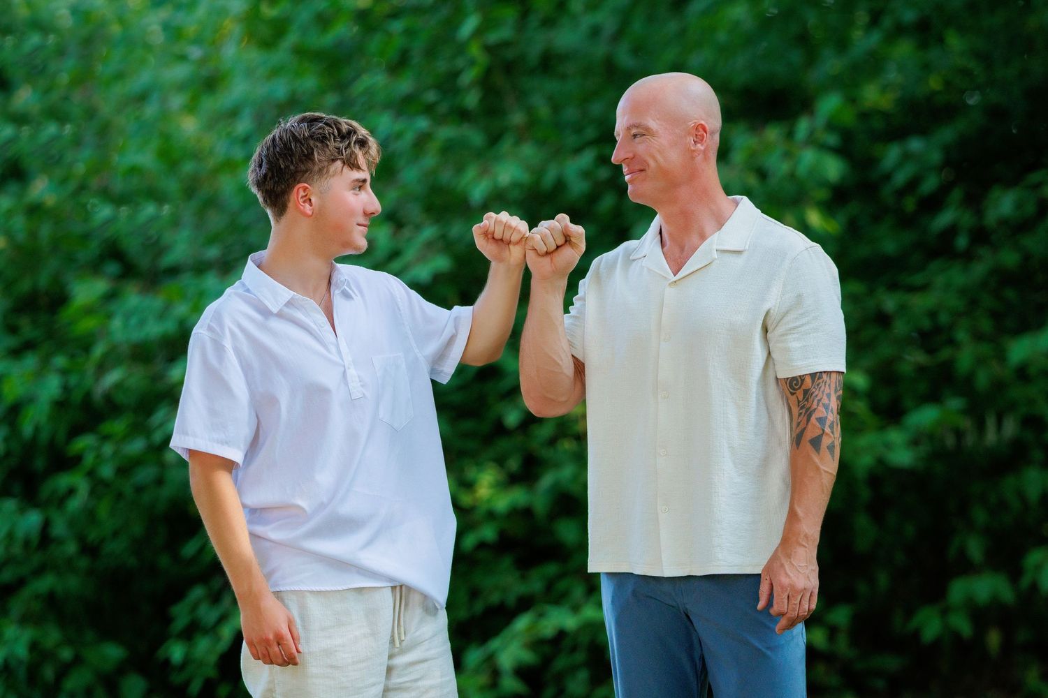 A bald tattooed man and teenage boy share a fist bump outdoors surrounded by green trees.