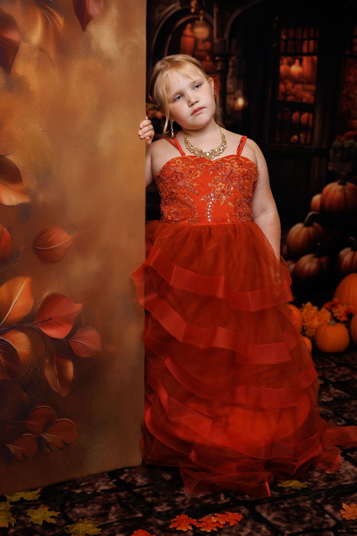 Young girl in elegant red ruffled gown poses beside autumn backdrop with pumpkins and fall leaves.