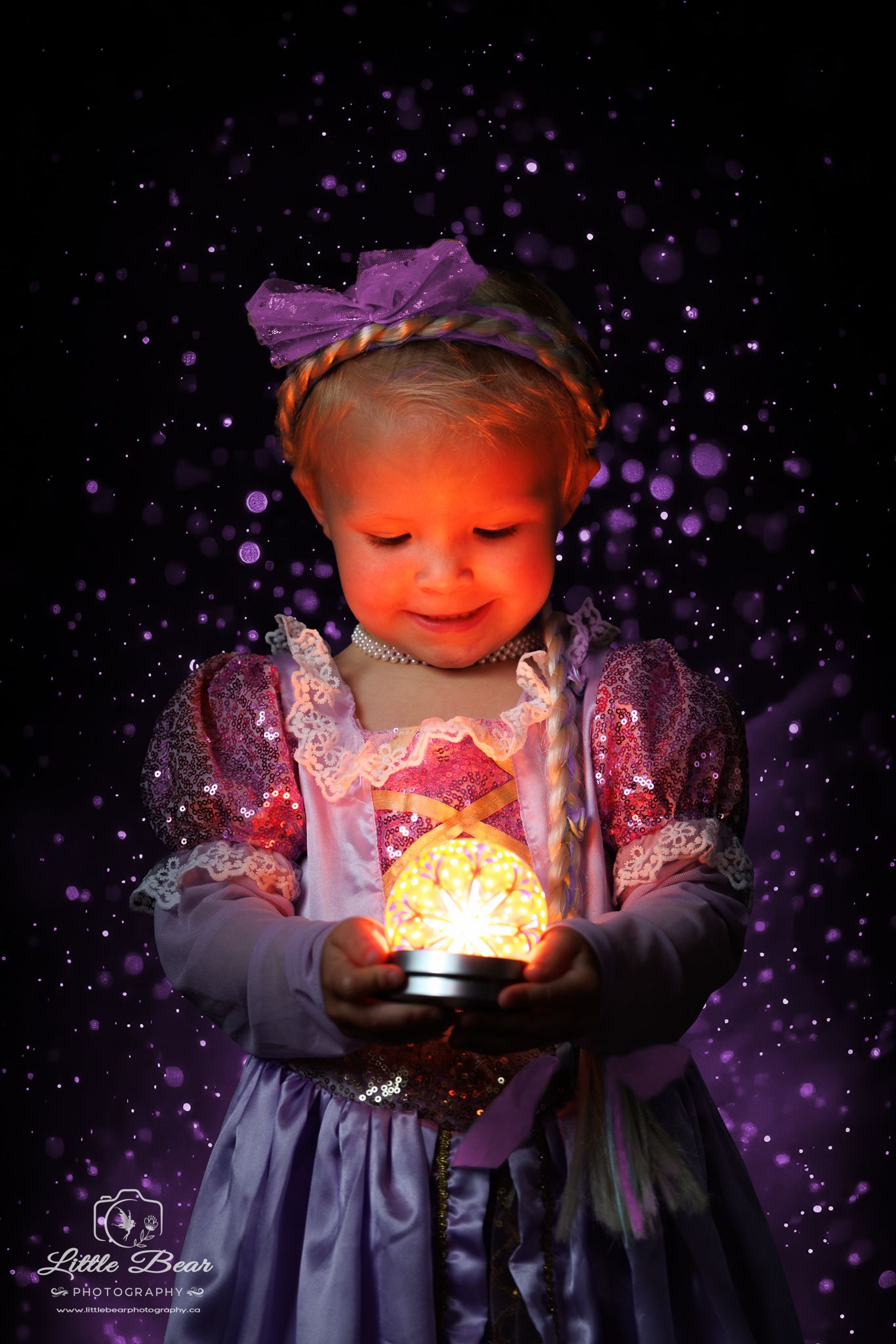 A toddler girl in a Rapunzel princess costume holds a glowing magical orb with purple bokeh sparkles in the background.