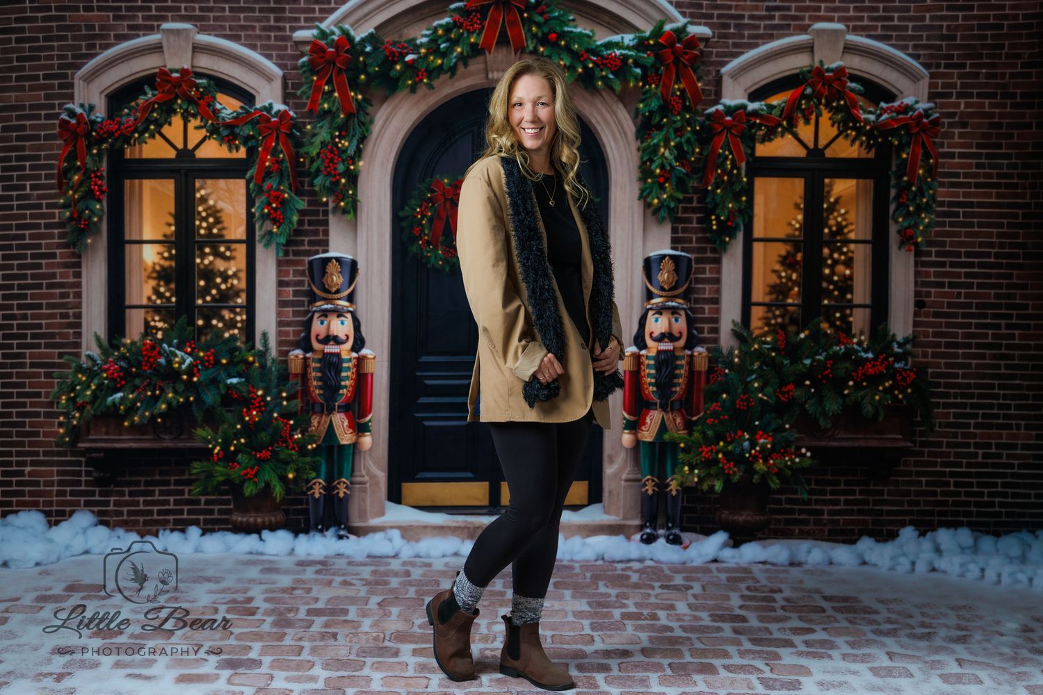 Woman in tan coat poses before festive brick building with nutcrackers, Christmas lights, and wreath-adorned door in snow.