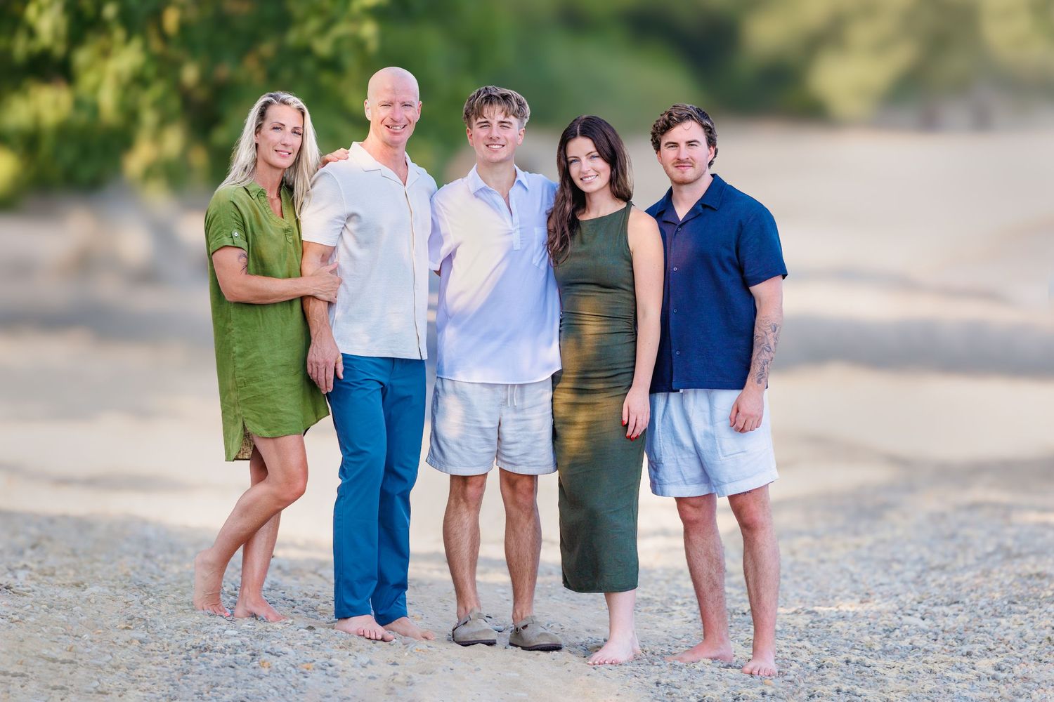 Family of five smiling together on a sandy beach, dressed in coordinated green, white, and blue outfits.