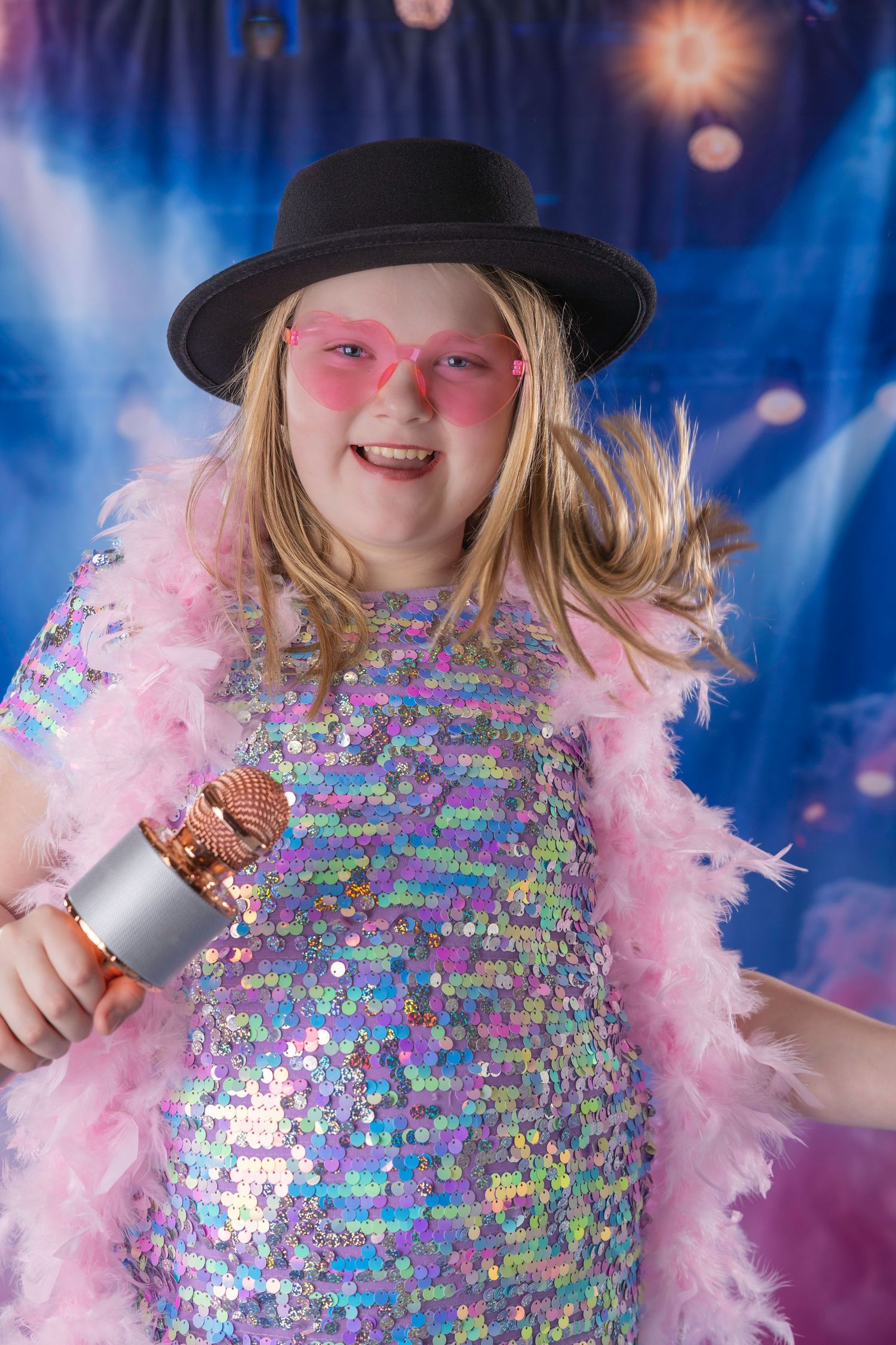 Energetic girl in sequin dress, black hat, and pink feather boa holds microphone on vibrant blue stage.