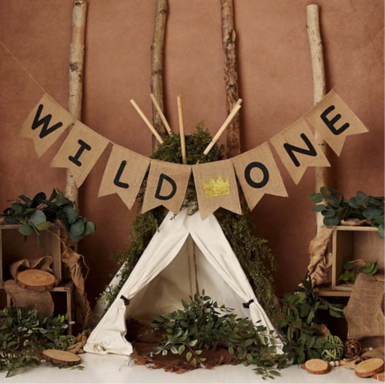 Rustic Wild One birthday party backdrop with burlap banner, teepee, greenery, and wooden accents on terracotta background.