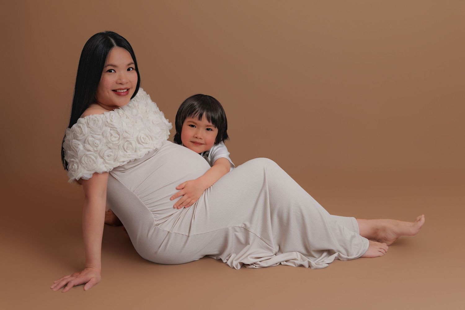 Pregnant woman in white dress poses with young child against warm beige background in maternity photo.