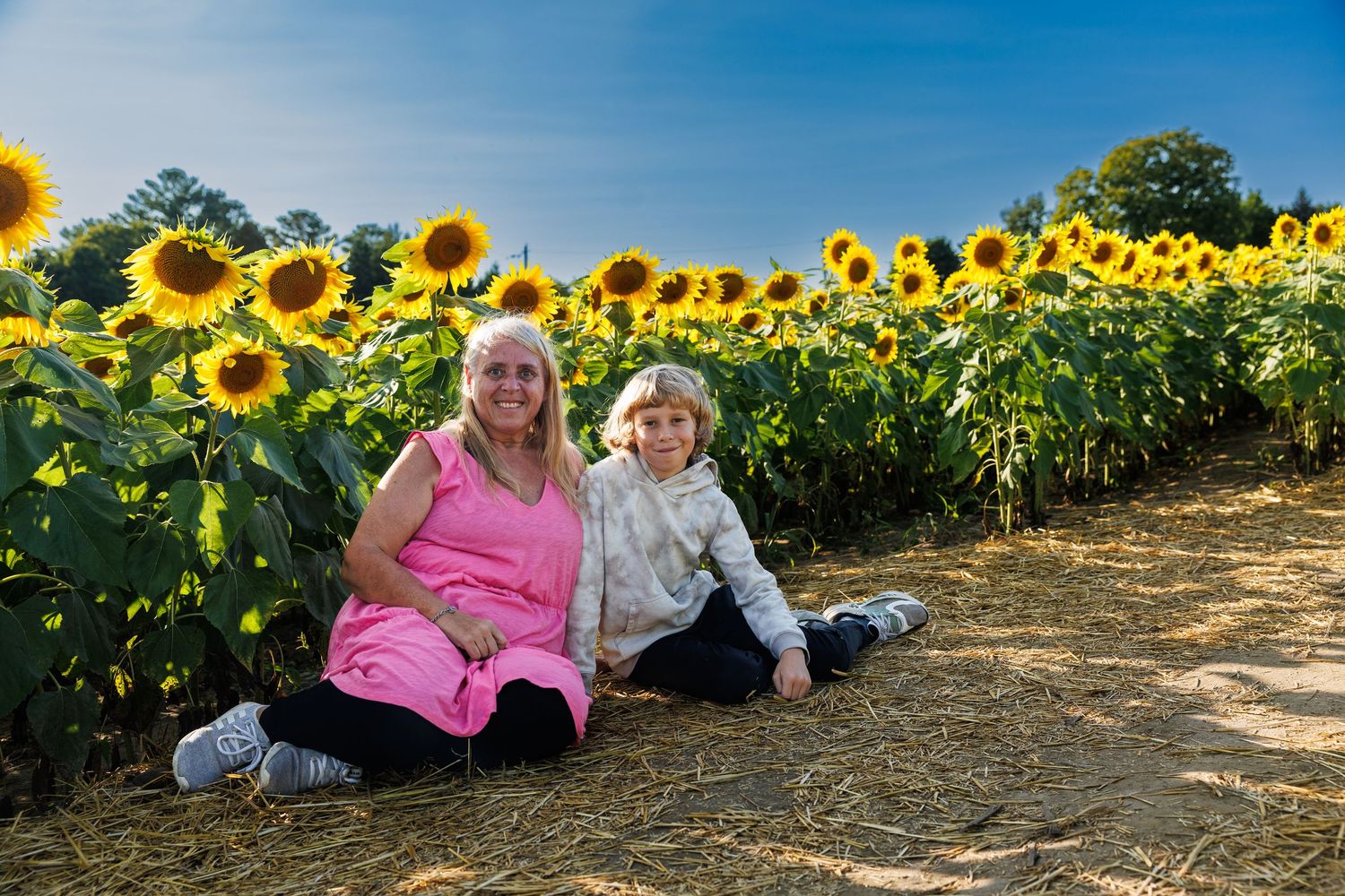 Woman and child sitting on hay in front of a bright sunflower field on a sunny day.