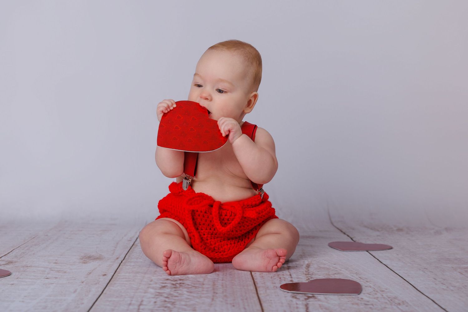 Adorable baby in red ruffled bloomers sits on wooden floor chewing a red heart-shaped toy.