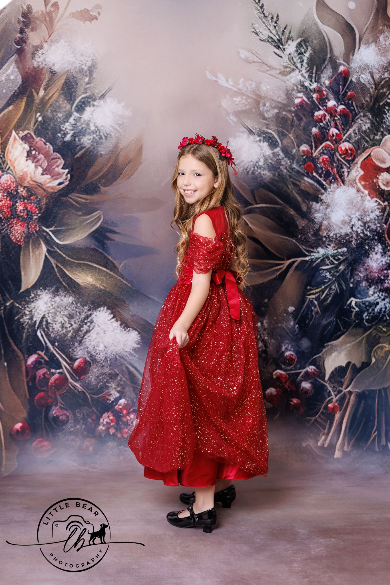 A young girl in a sparkling red gown and floral crown poses before a festive winter floral backdrop.