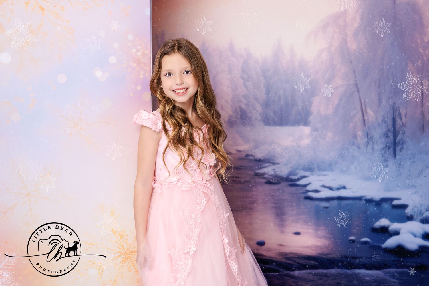 A young girl in a pink ball gown poses gracefully before a dreamy winter wonderland backdrop with snow and a stream.